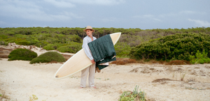 Ein Mann mit Surfboard im Arm, über dem eine Decke hängt, steht am Strand