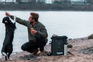 Mann mit Getränk in der Hand spielt mit einem Hund Stöckchen, neben ihm steht ein Kühlrucksack und Thermoflasche im Sand